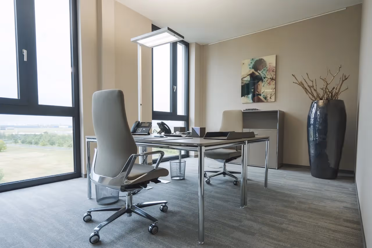 Helles Büro mit zwei Stühlen, großem Fenster, modernem Tisch, Kunstwerk an der Wand und großer Vase mit Ästen.