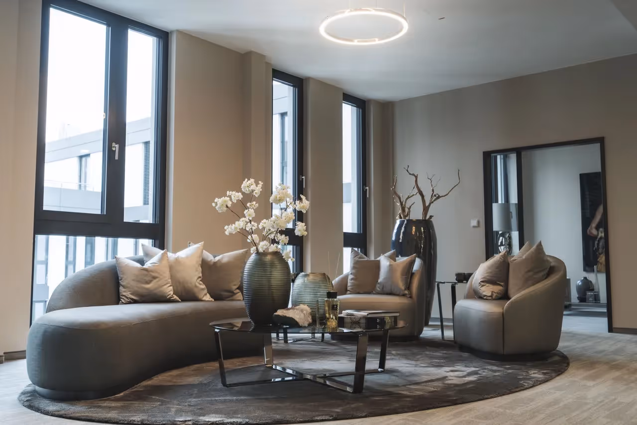 Ecksofa mit Kissen, runder Tisch, Blumenvasen, große Fenster, runde Deckenleuchte, grauer Teppich, moderne Einrichtung.