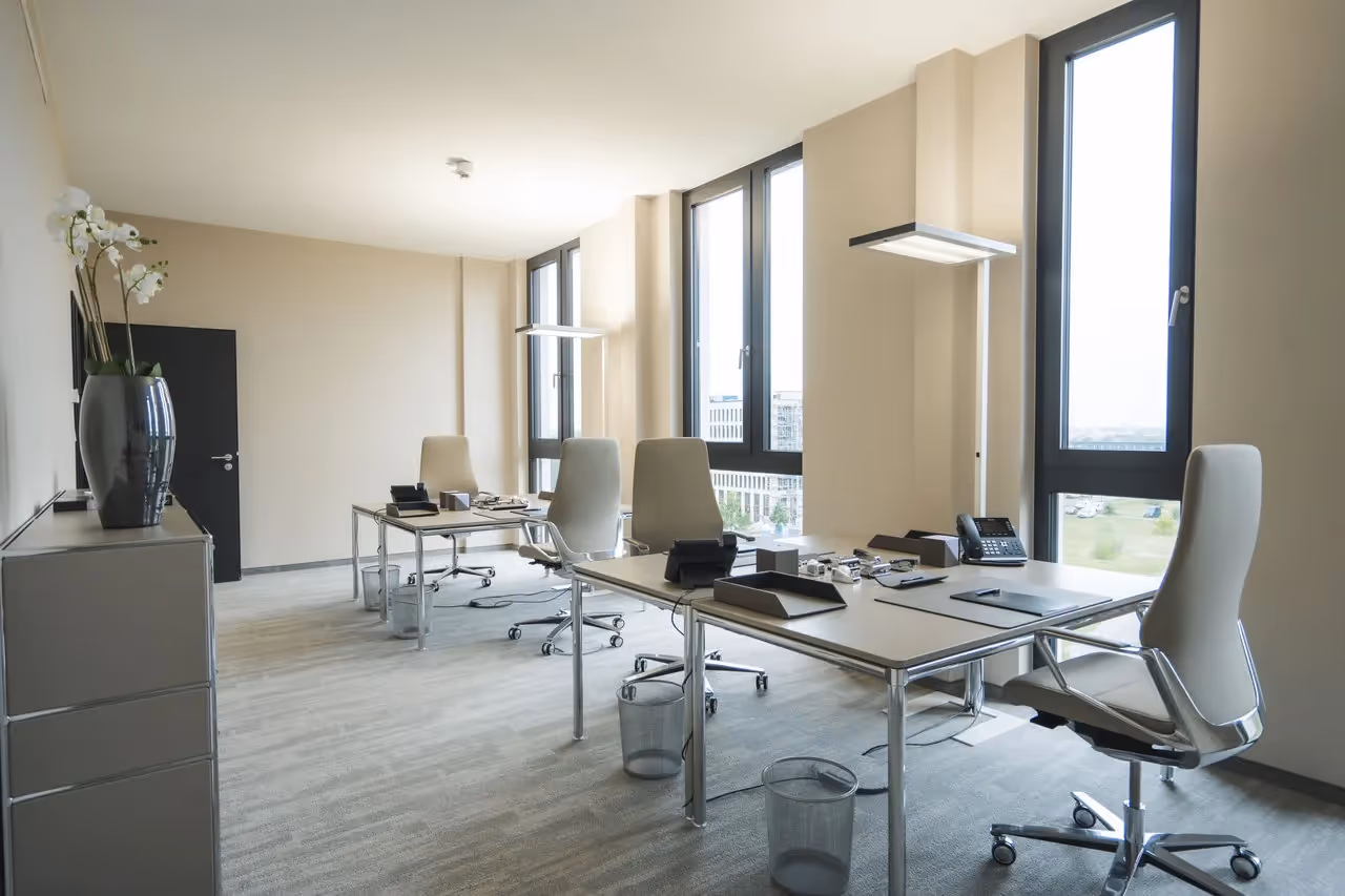 Helles Büro mit vier Schreibtischen, modernen Stühlen, großen Fenstern und grauem Teppichboden.