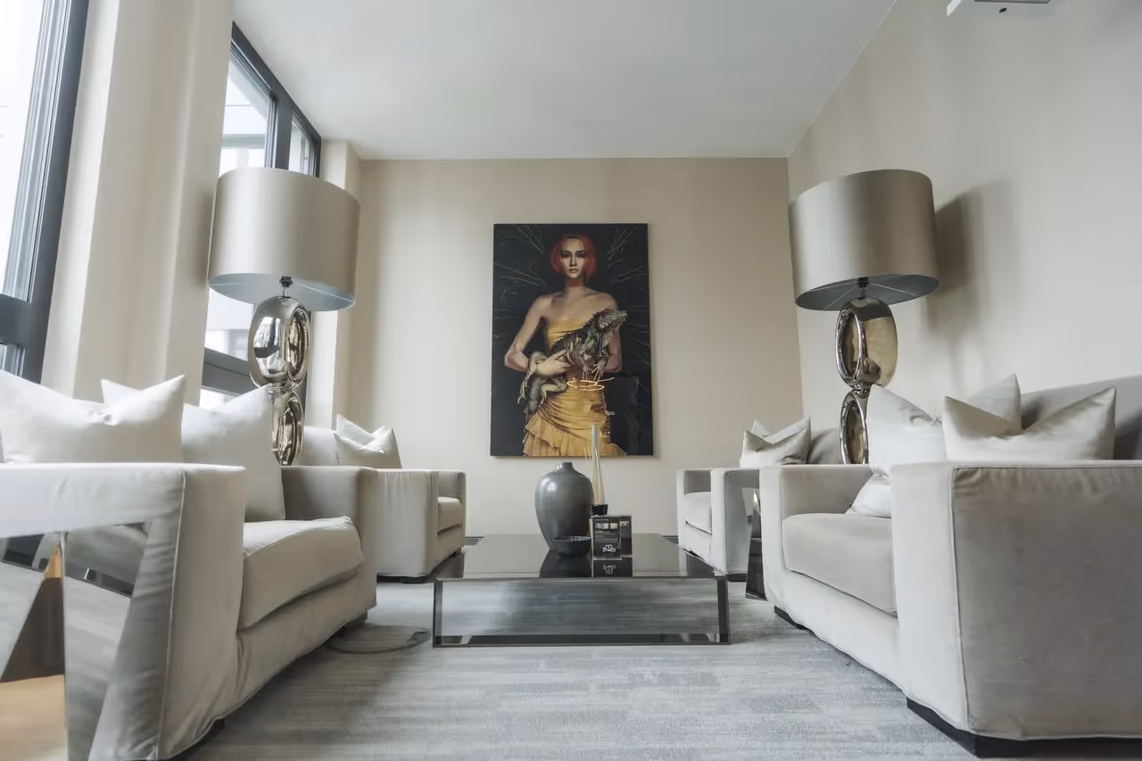 Zwei beige Sofas mit Kissen, Glastisch, große Lampen, Wandbild einer Frau, grauer Teppich, große Fenster.