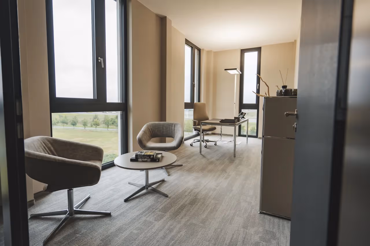Helles Büro mit zwei Sesseln, rundem Tisch, Schreibtisch, großen Fenstern und Teppichboden.