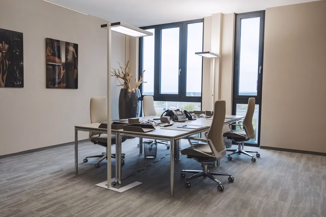 Moderner Besprechungsraum mit vier Bürostühlen, großem Tisch, Stehlampen, großen Fenstern und Wandbildern.