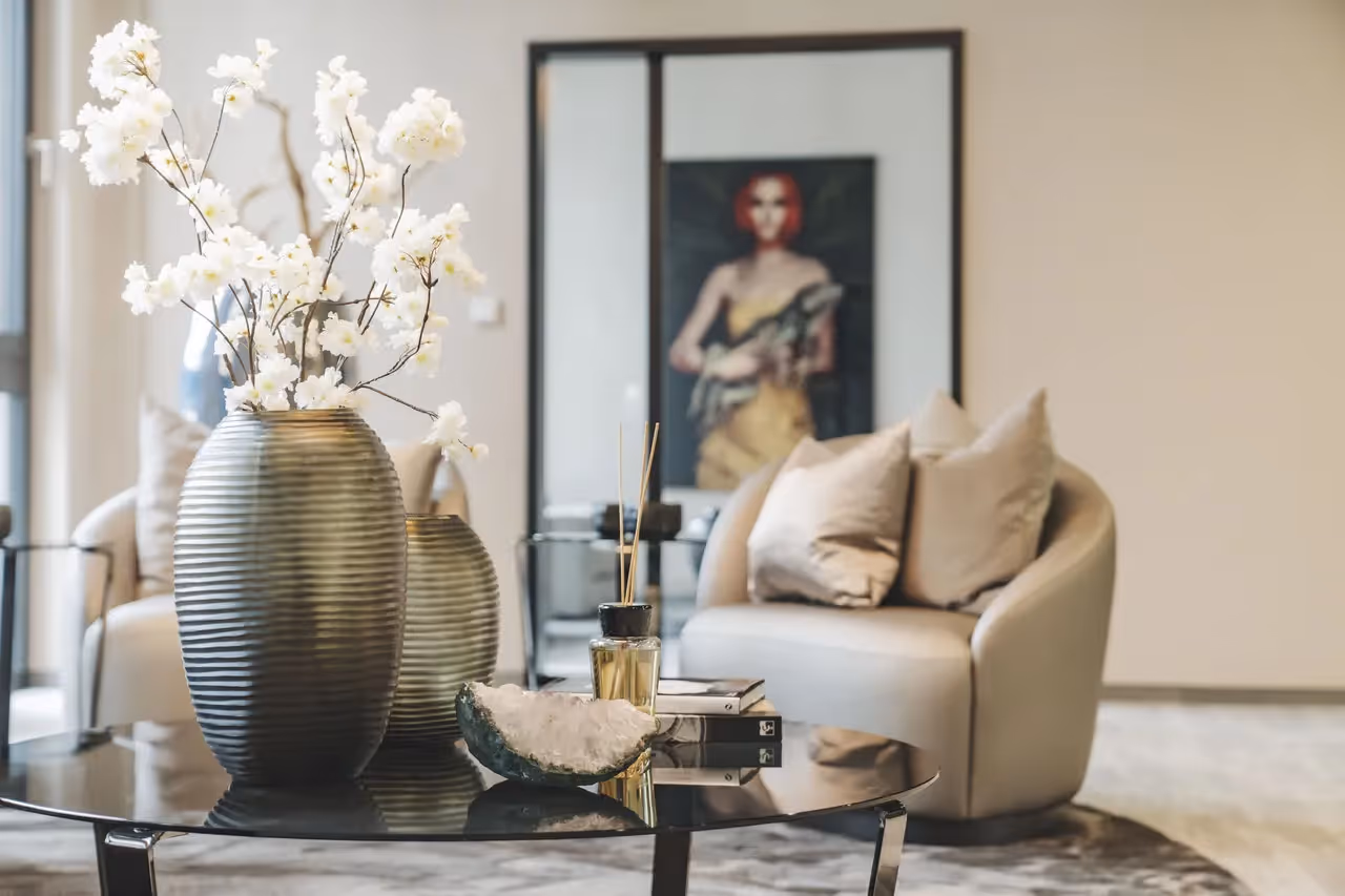Runder Glastisch mit Vase und Deko vor beigem Sessel, Kunstwerk im Hintergrund.
