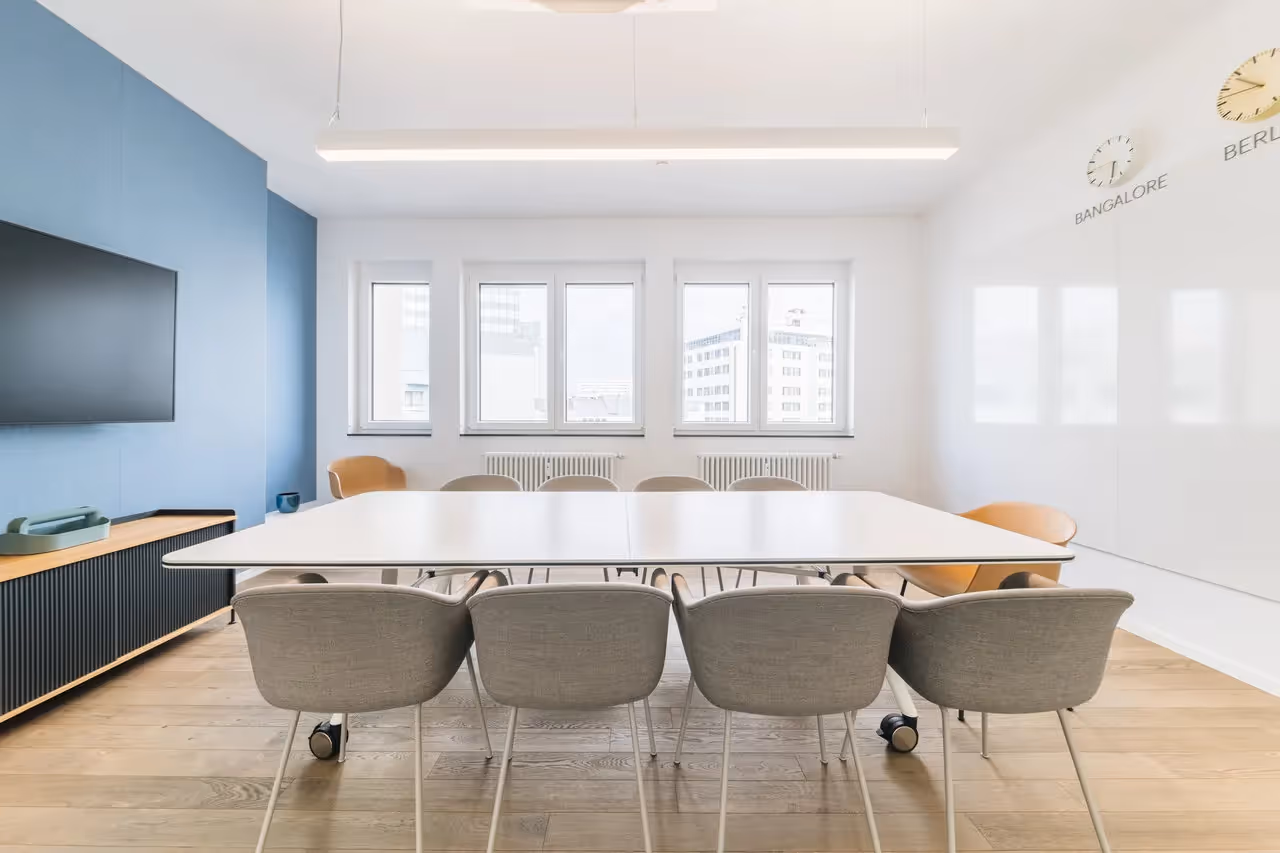 Heller Konferenzraum mit großem Tisch, grauen Stühlen, blauer Wand, Wanduhren und Fenstern mit Stadtblick.
