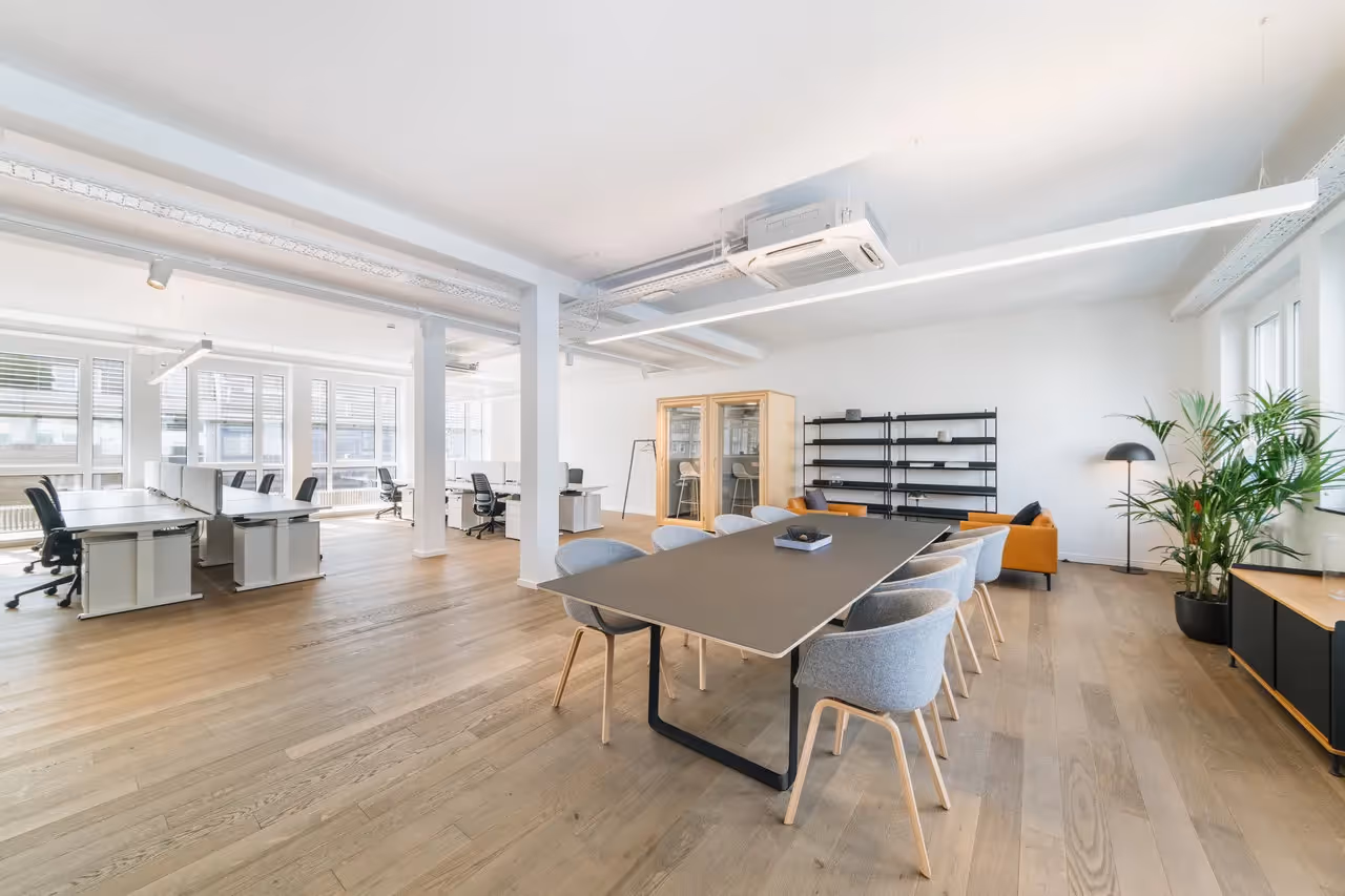 Heller Raum mit Holzboden, großem Tisch, grauen Stühlen, Regalen, Pflanzen, Sofaecke und Schreibtischen.