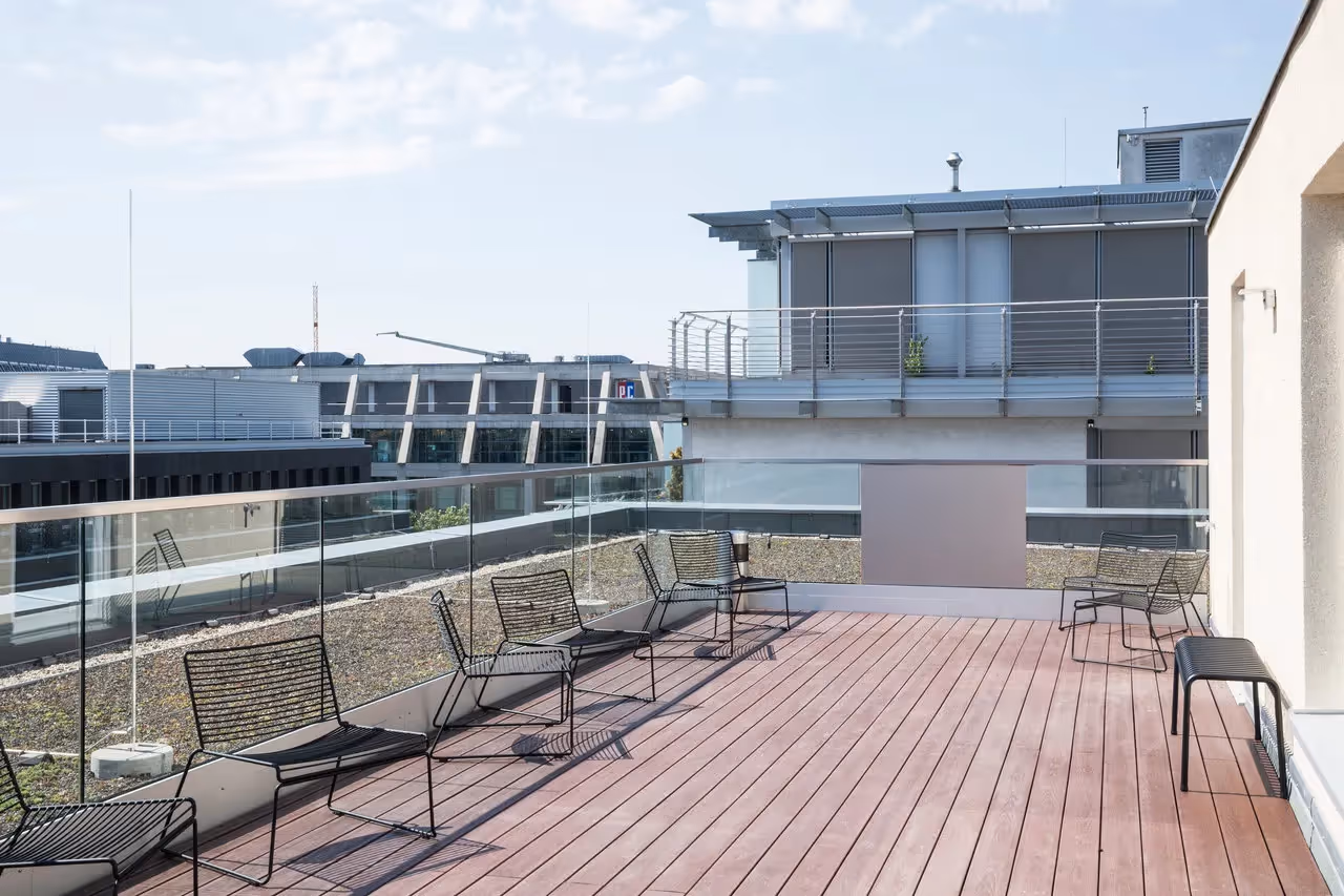 Dachterrasse mit Holzboden, Glasgeländer, schwarzen Metallstühlen, Blick auf modernes Gebäude bei klarem Himmel.