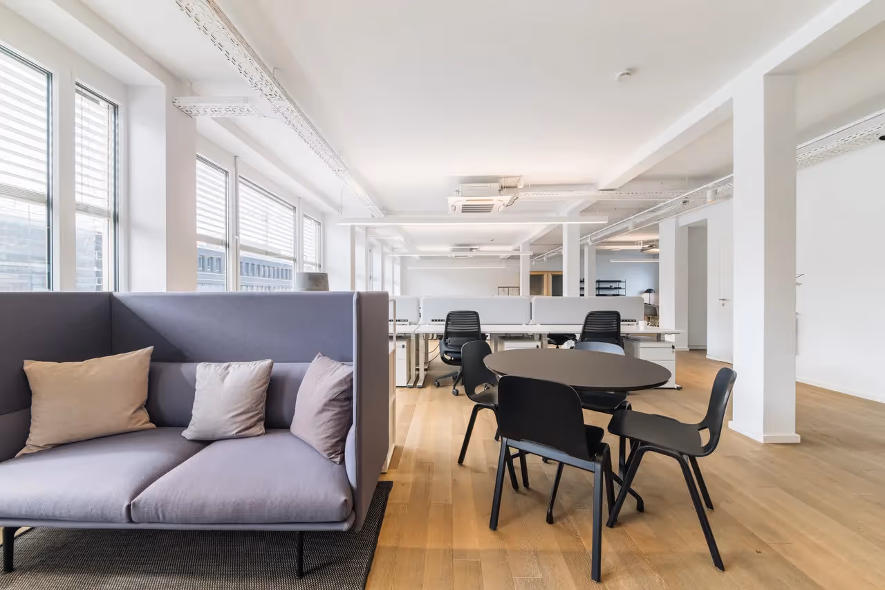 Heller Raum mit lila Sofa, Holzboden, runden Tisch mit schwarzen Stühlen, großen Fenstern und weißen Wänden.