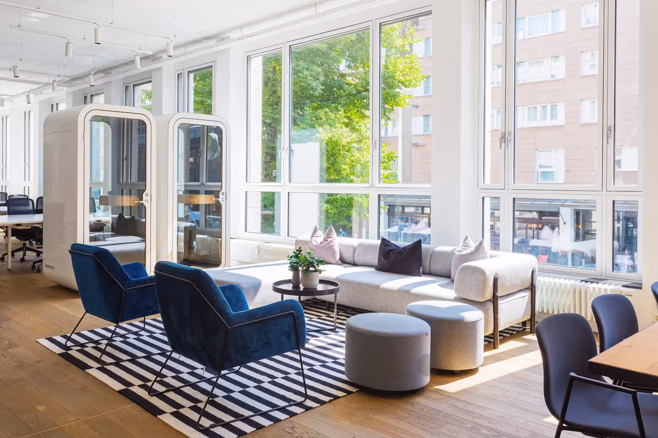 Heller Raum mit großen Fenstern, grauem Sofa, blauen Sesseln, Streifenteppich, Holzboden, zwei Telefonboxen.