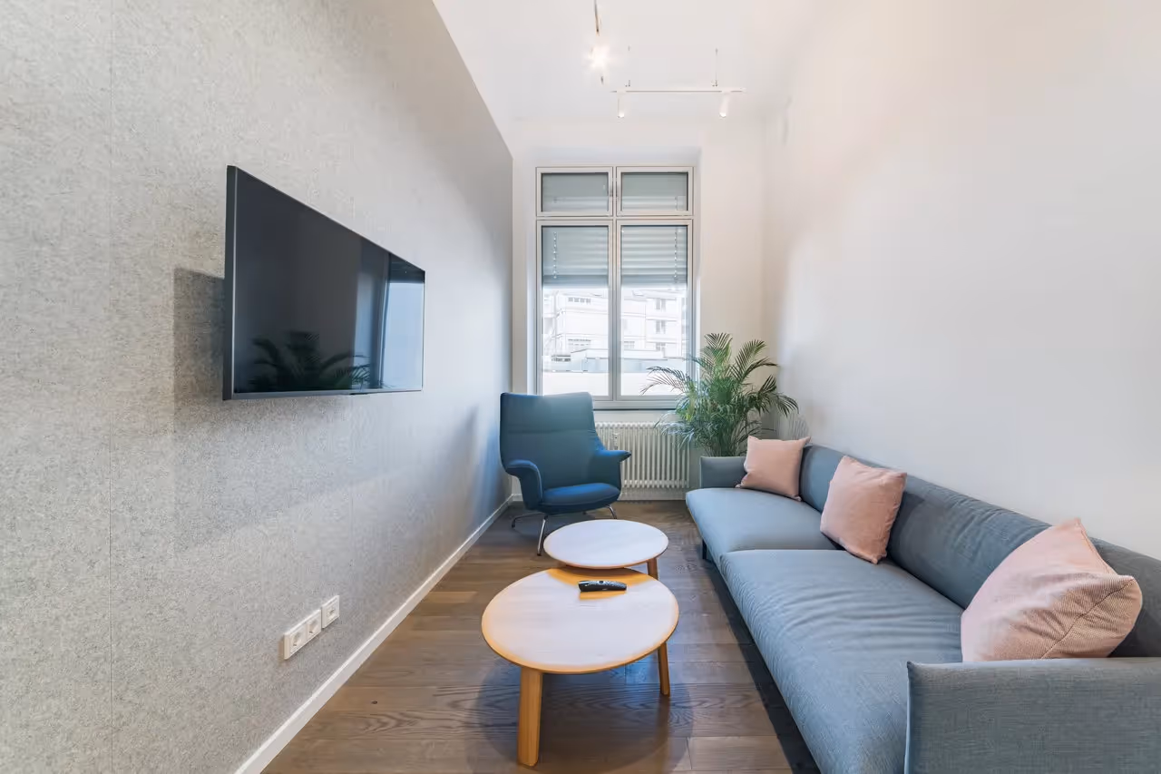 Graues Sofa, blauer Sessel, Holztische, Wand-TV, große Fenster, Pflanzen, Holzfußboden, helle Beleuchtung.