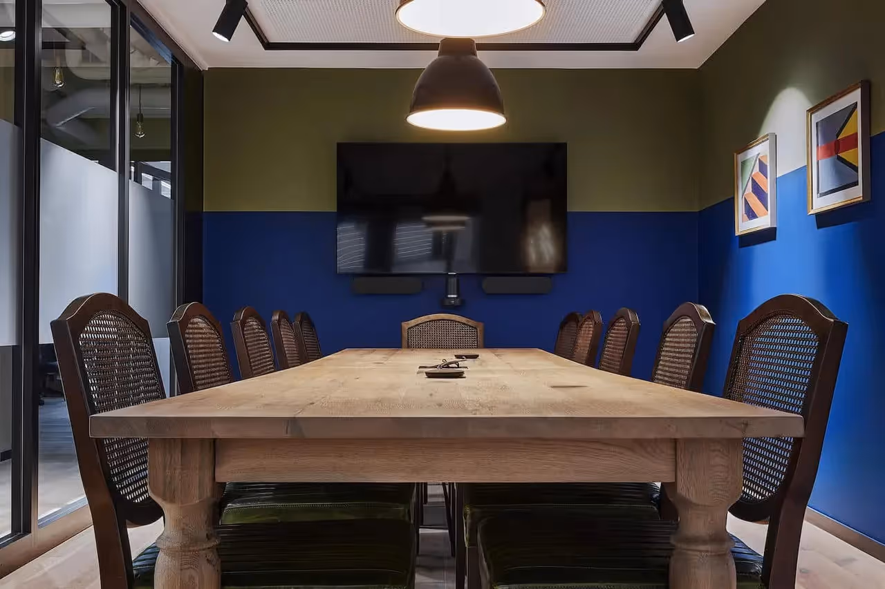 Großer Holztisch mit geflochtenen Stühlen, blauer und grüner Wand, Deckenleuchte, TV-Bildschirm, Glaswände.