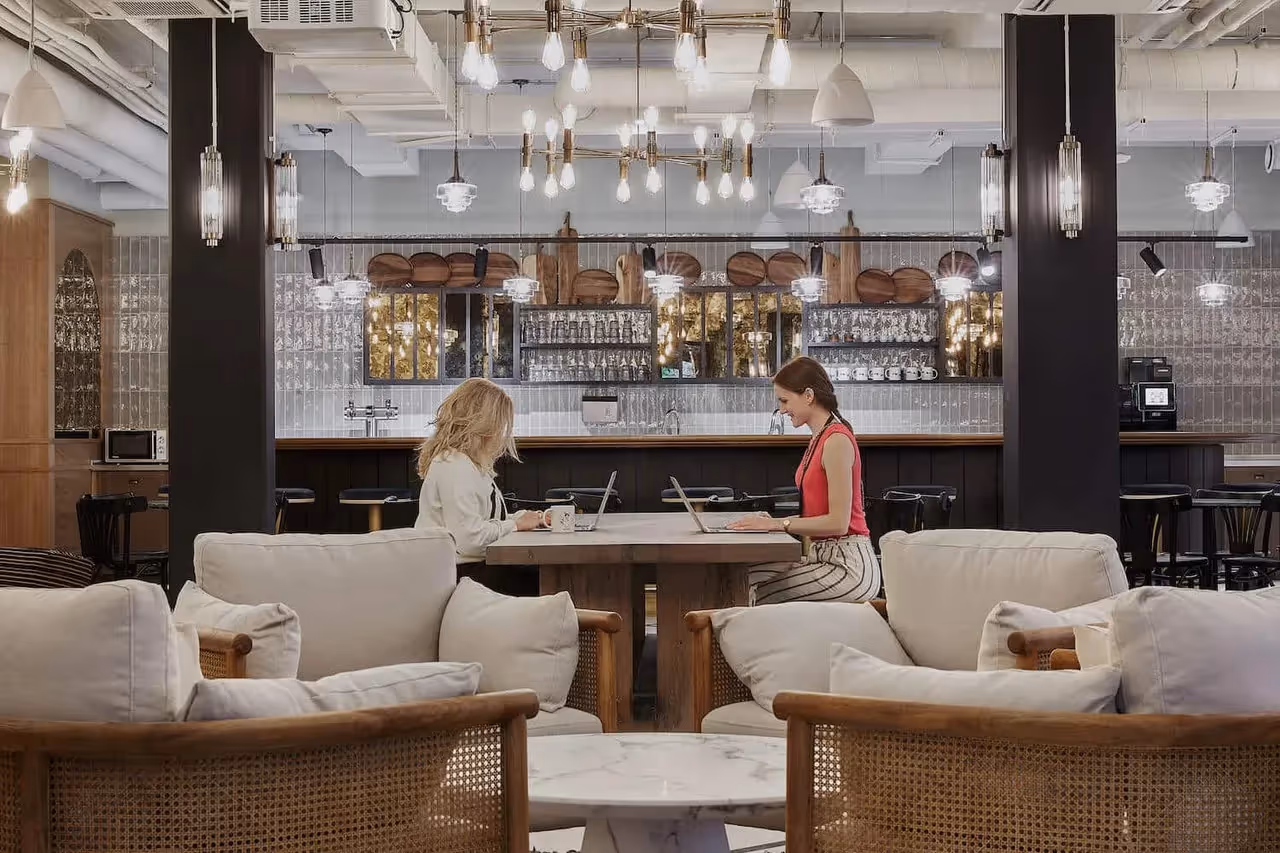Zwei Personen arbeiten an einem Holztisch, umgeben von Sofas und moderner Beleuchtung, Bar im Hintergrund.