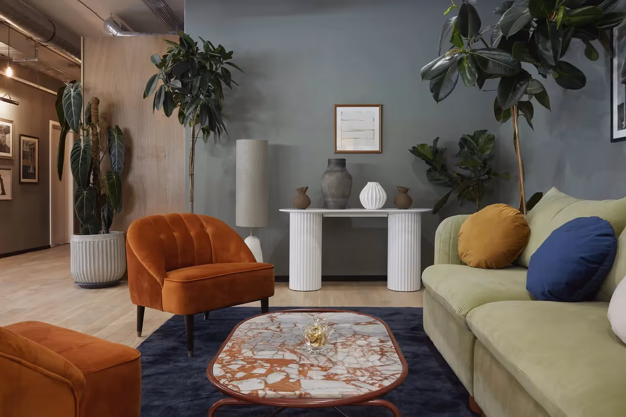Grünes Sofa, orange Sessel, Marmortisch, Pflanzen, weiße Konsole mit Vasen, Wandbild, Holzfußboden, Stehlampe.