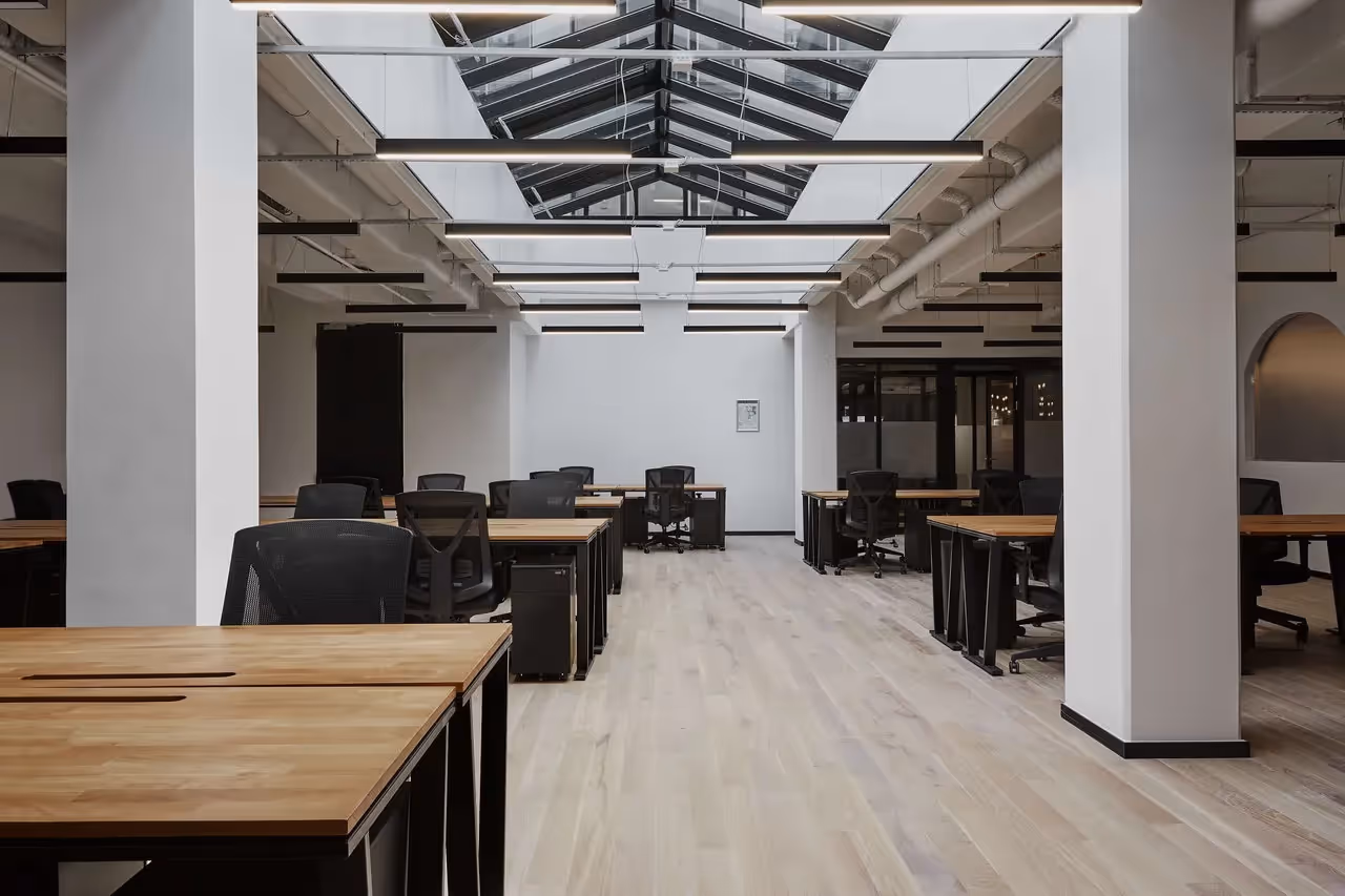 Großer Raum mit Holztischen, schwarzen Bürostühlen, Oberlichtern und hellen Holzböden in einem Coworking-Space.