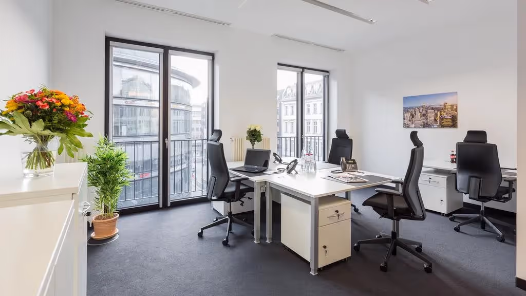 Helles Büro mit vier Schreibtischen, schwarzen Stühlen, großen Fenstern, Blumen und Stadtbild an der Wand.