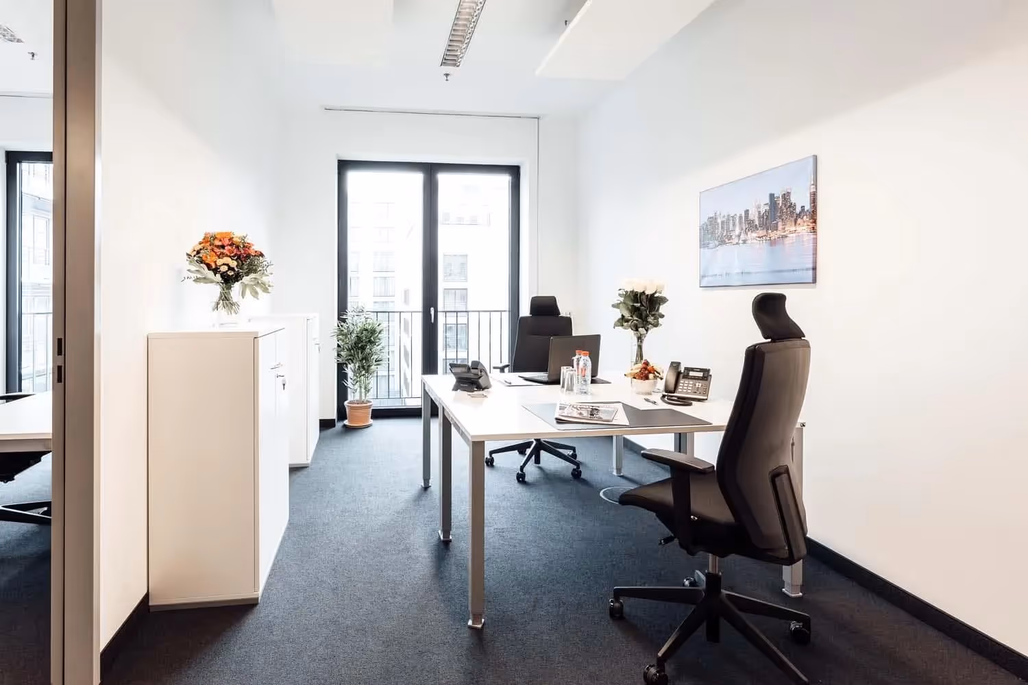 Helles Büro mit zwei Schreibtischen, schwarzen Bürostühlen, Blumen, großem Fenster, Teppichboden und Wandbild.