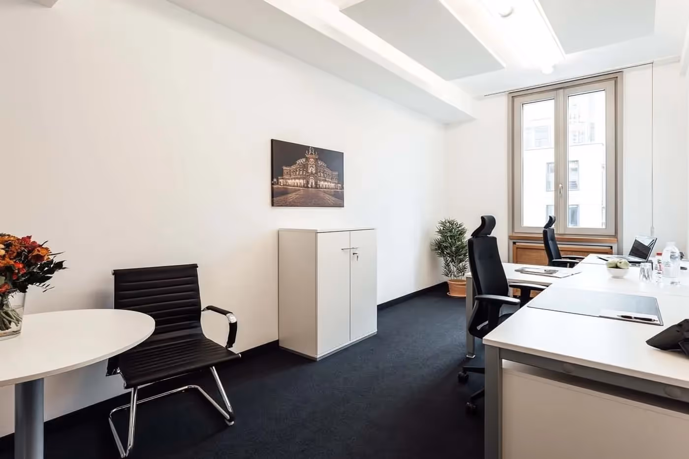 Helles Büro mit zwei Schreibtischen, schwarzen Stühlen, großem Fenster, Pflanzen, weißem Schrank und Bild an der Wand.