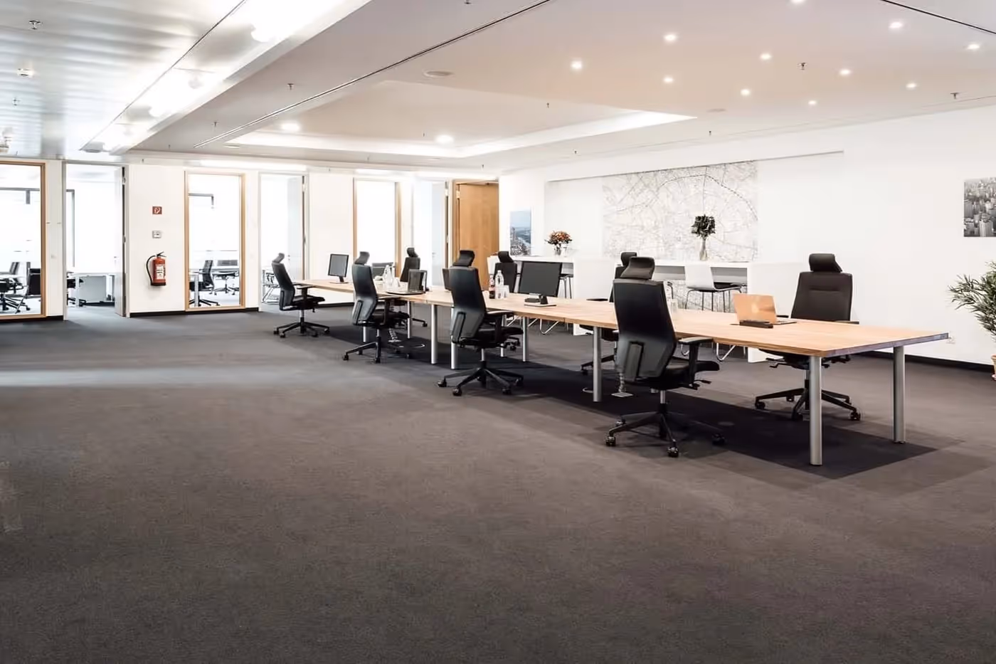 Großer, heller Raum mit langen Holztischen, schwarzen Bürostühlen, Teppichboden und moderner Deckenbeleuchtung.