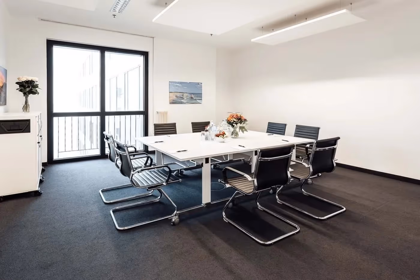 Konferenzraum mit großem Tisch, schwarzen Stühlen, Blumenstrauß, großem Fenster, weißem Schrank und Deckenbeleuchtung.