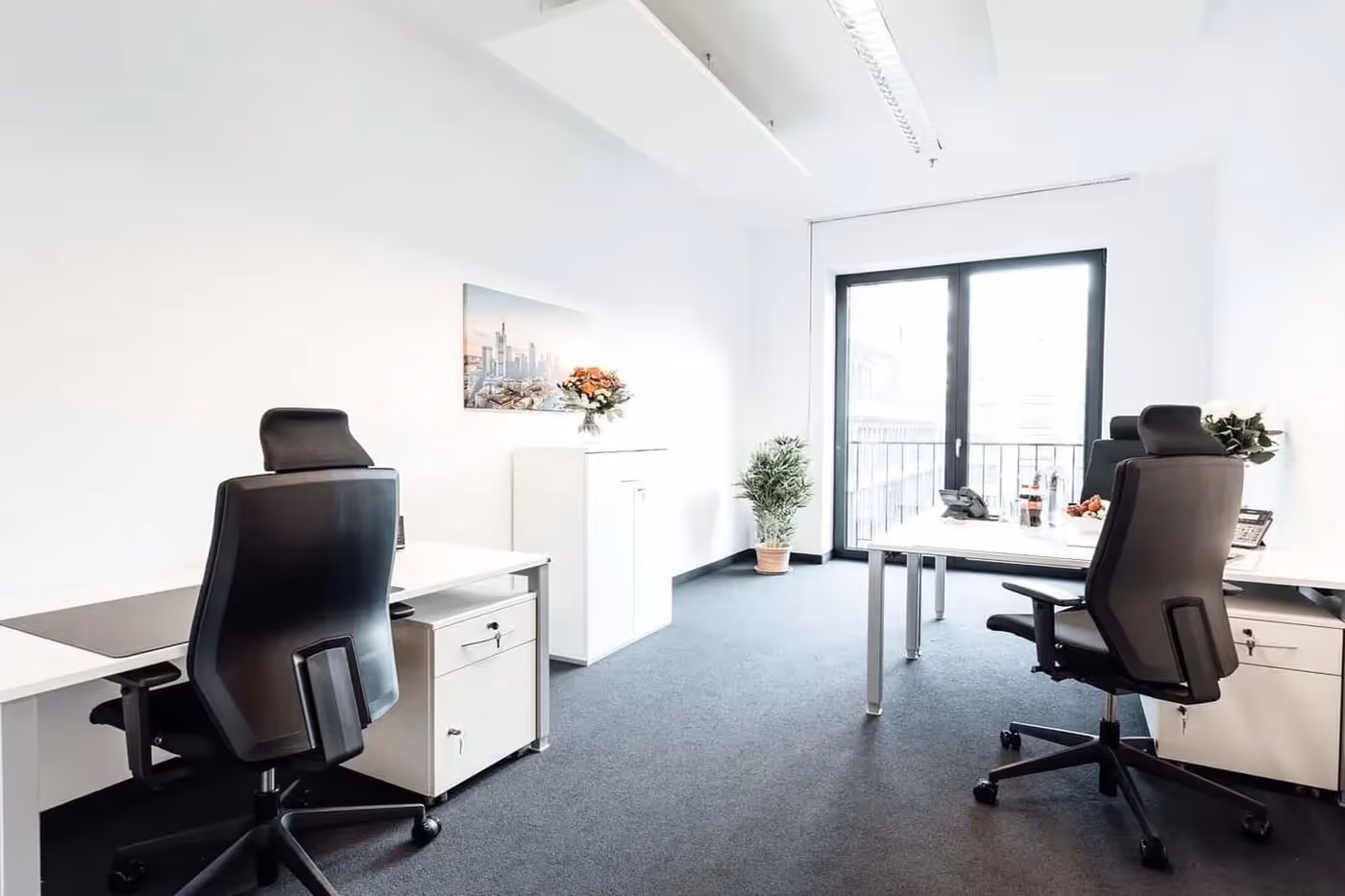 Helles Büro mit zwei Schreibtischen, schwarzen Stühlen, großem Fenster, Pflanzen und Wandbild.