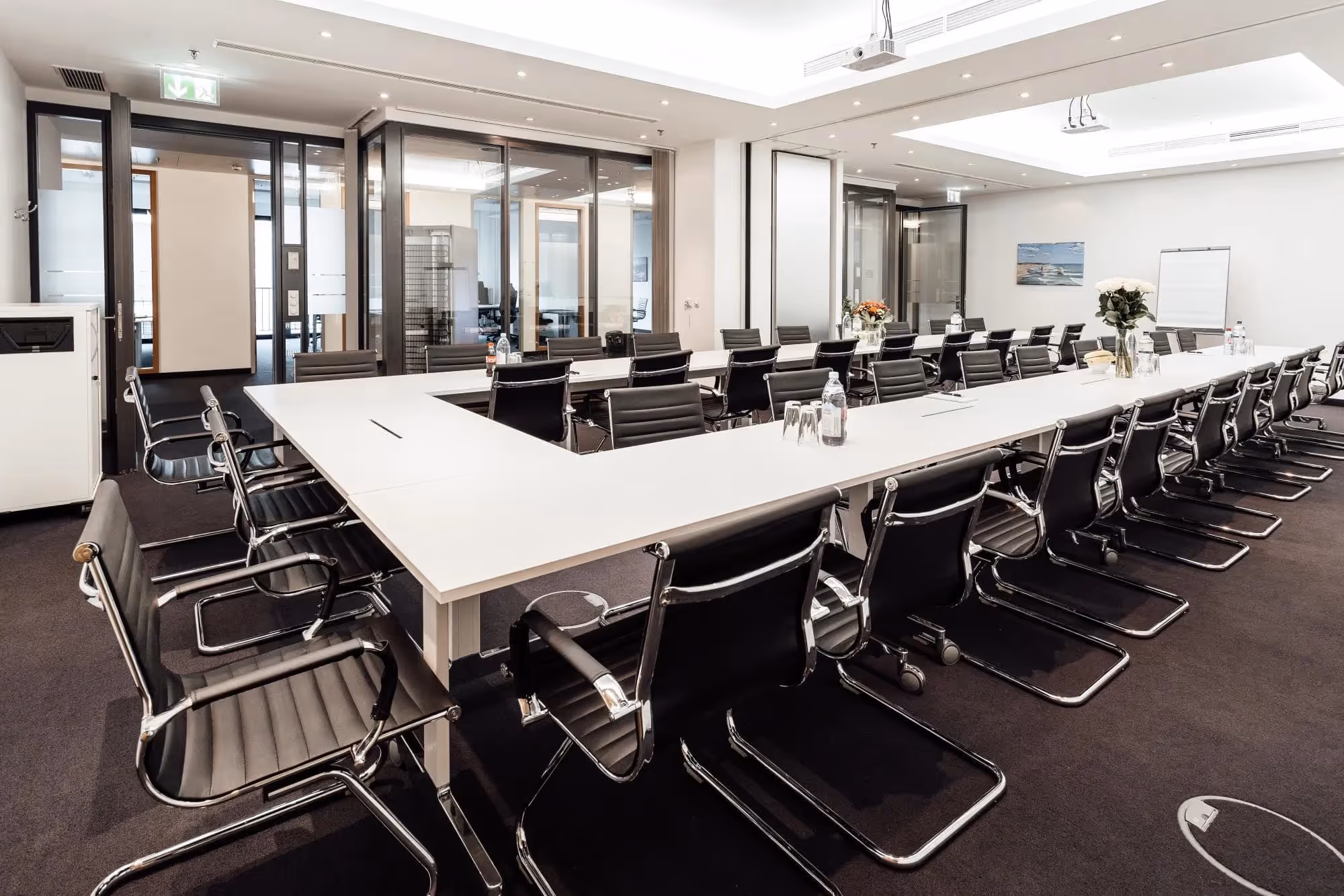 Konferenzraum mit U-förmigem Tisch, schwarzen Lederstühlen, Glaswänden, Deckenleuchten und Blumenarrangement.
