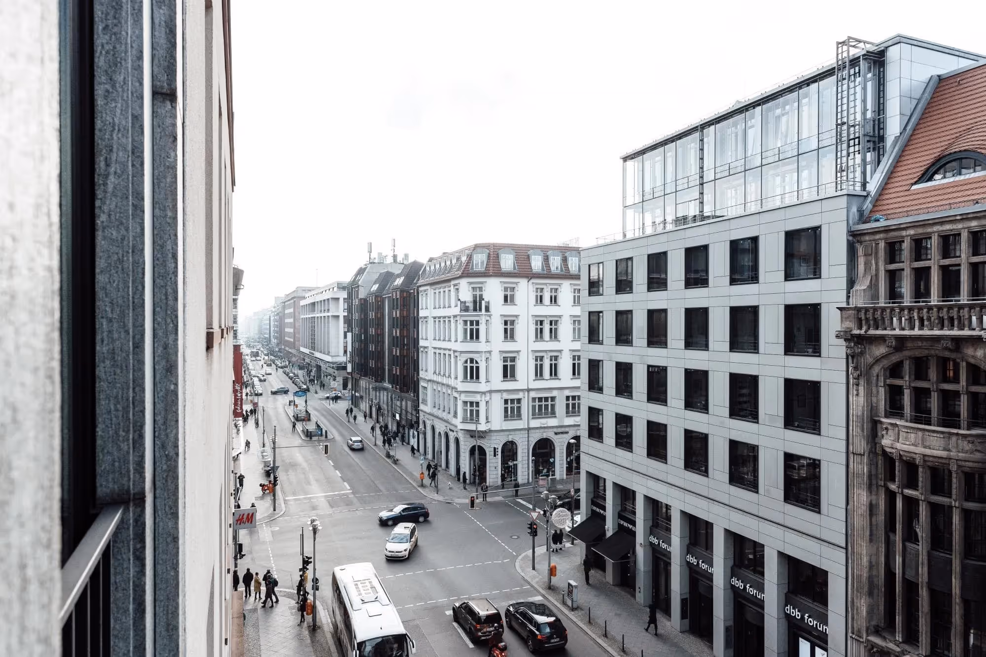 Straßenansicht mit historischen und modernen Gebäuden, Fußgängern und Autos bei bewölktem Himmel.