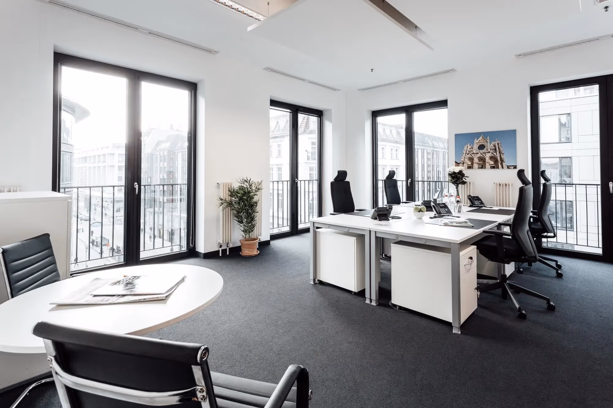 Helles Büro mit großen Fenstern, weißen Schreibtischen, schwarzen Stühlen und einer Pflanze in der Ecke.
