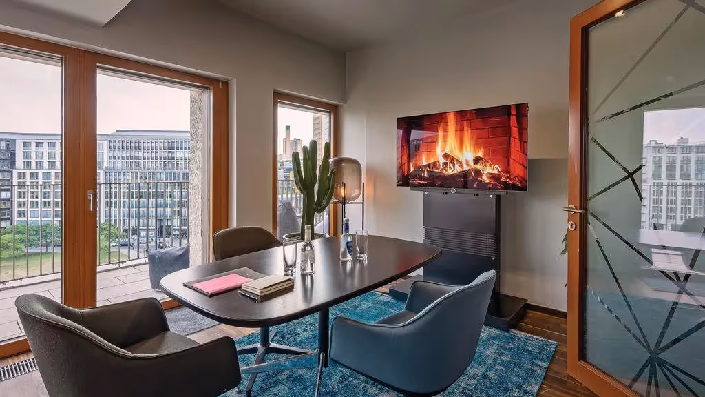 Konferenzraum mit ovalem Tisch, vier Stühlen, TV mit Kaminbild, großen Fenstern, Balkonblick und blauem Teppich.