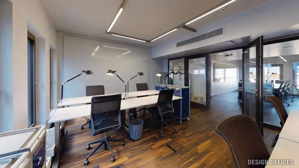 Heller Raum mit Holzboden, Schreibtischen, schwarzen Stühlen, Tischlampen, Glastüren und moderner Deckenbeleuchtung.