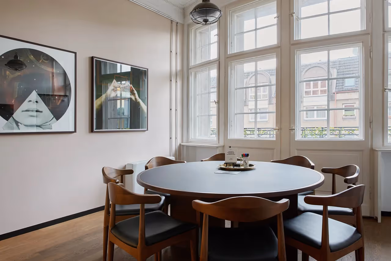 Runder Holztisch mit sechs Stühlen, helle Fenster, zwei moderne Kunstwerke an der Wand, Holzfußboden.