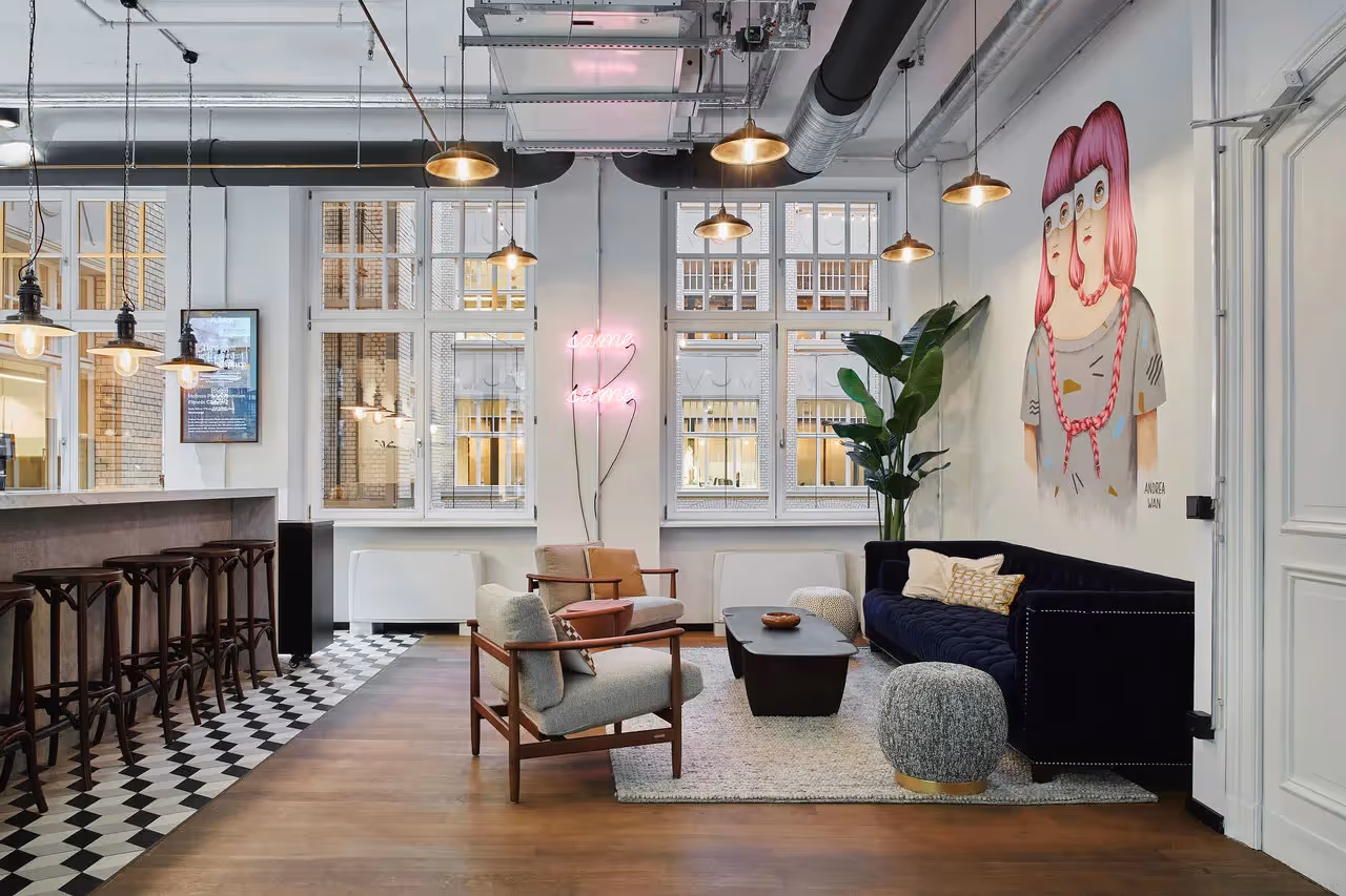 Loungebereich mit blauer Couch, grauem Teppich, Kunstwerk an der Wand, Barhockern und Hängelampen vor großen Fenstern.