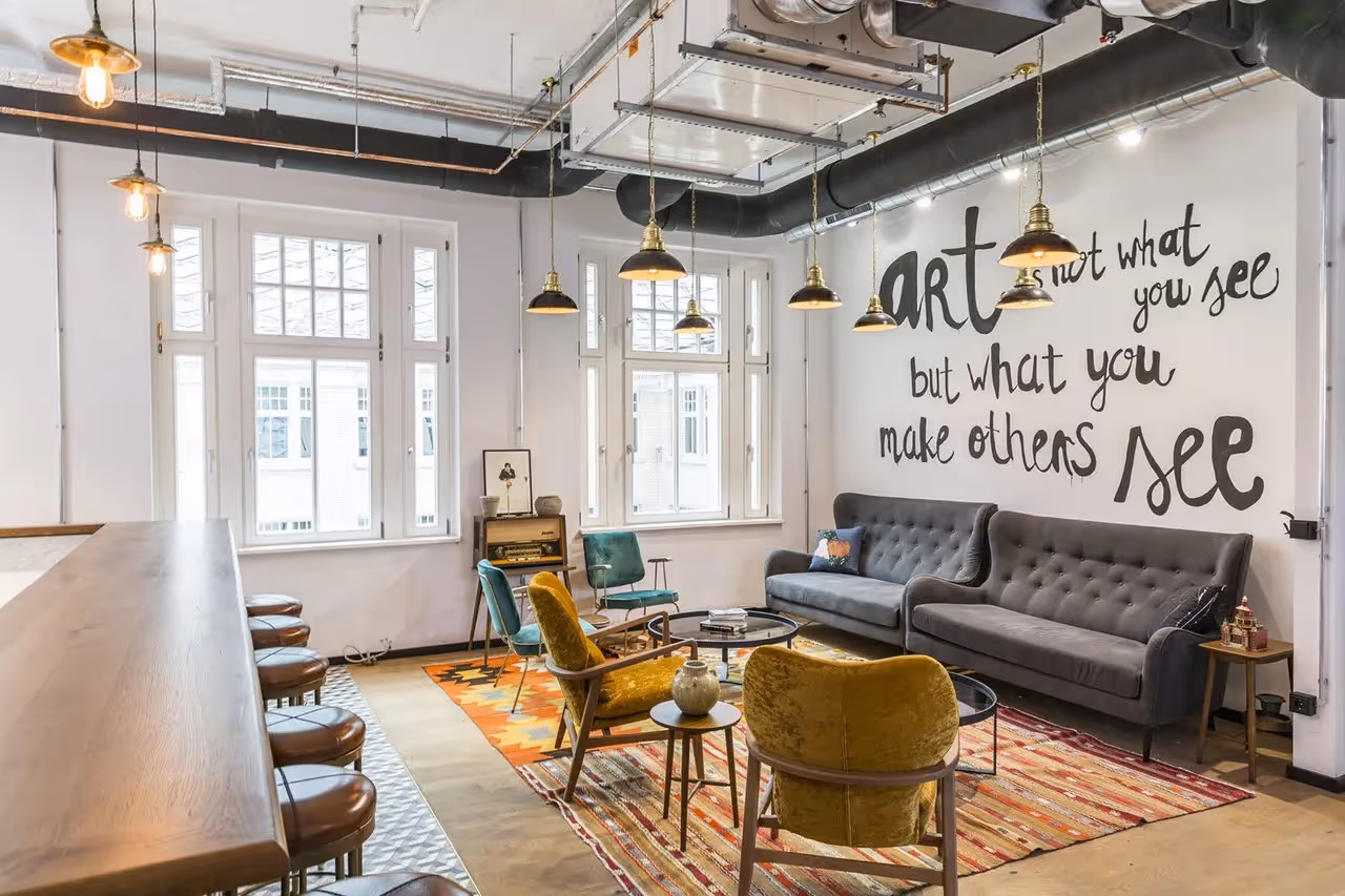 Gemütlicher Raum mit grauem Sofa, bunten Sesseln, Barhockern, Teppich, industriellen Hängelampen und Wandkunst.