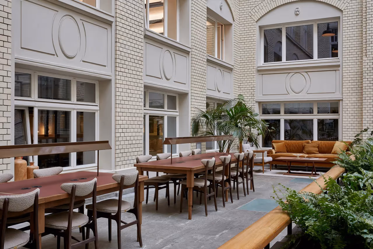 Heller Raum mit langen Holztischen, gepolsterten Stühlen, beigen Sofas, Pflanzen und großen Fenstern.