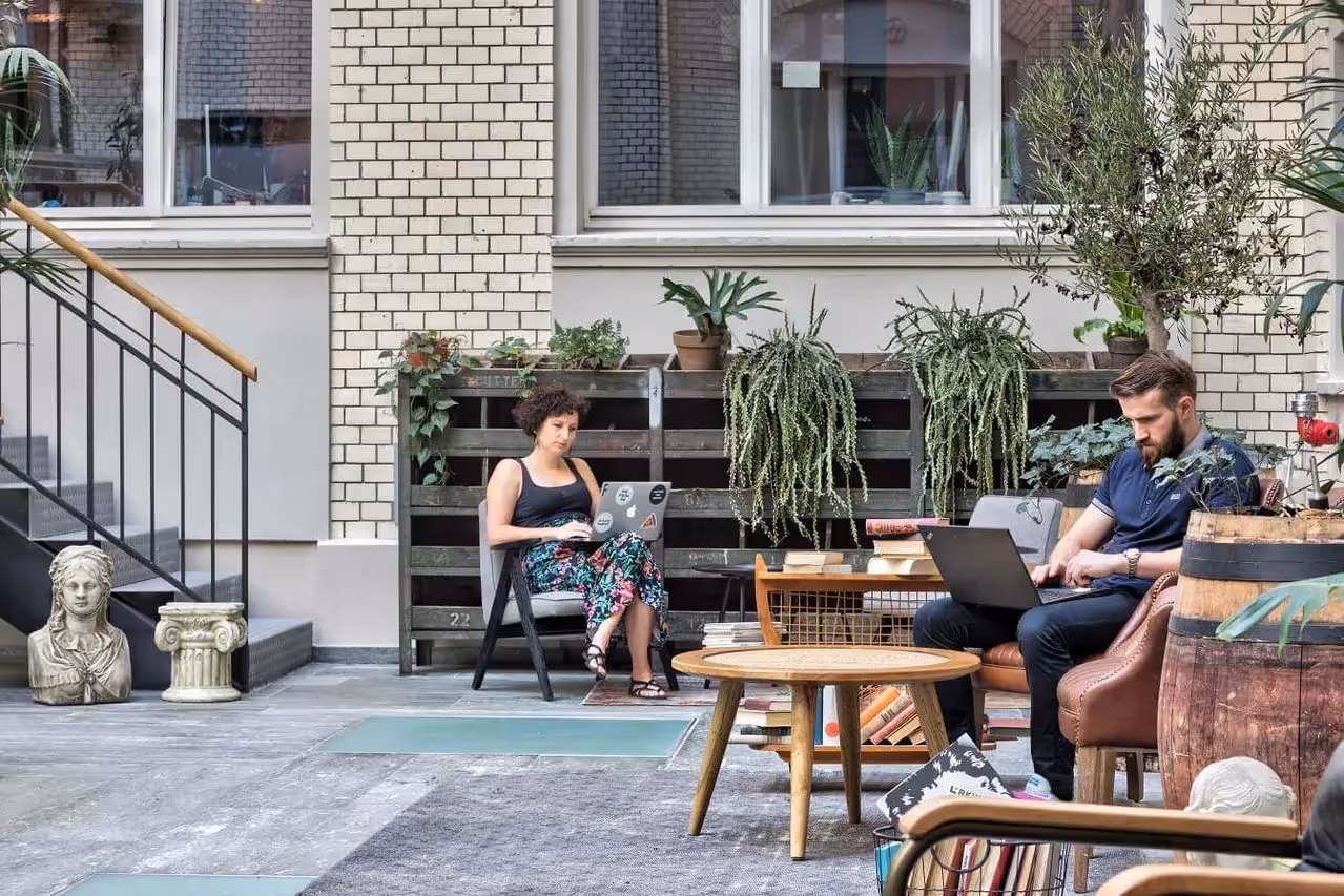 Zwei Personen arbeiten an Laptops, umgeben von Pflanzen, Holzmöbeln und einer Büste in einem begrünten Innenhof.