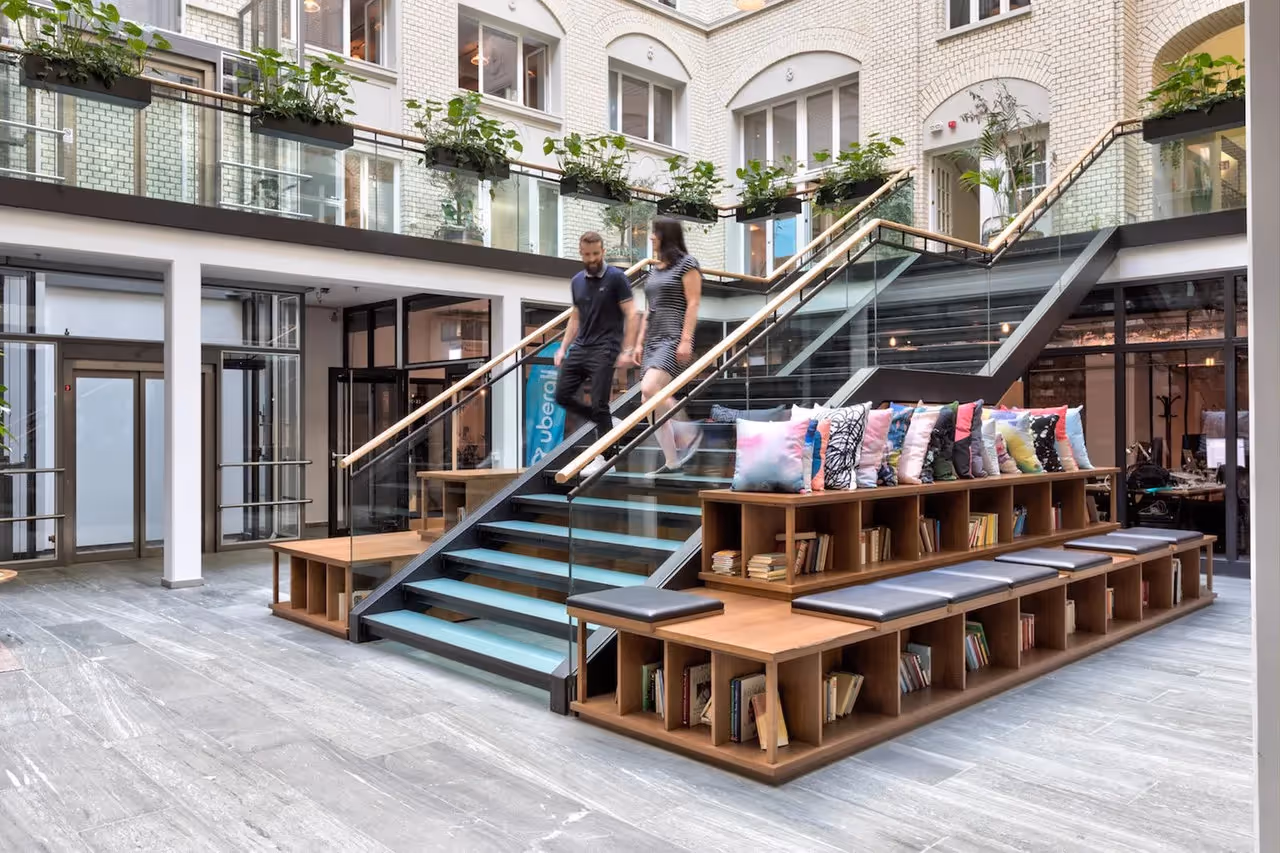 Glasgeländer-Treppe mit Holzsitzbank, bunten Kissen, Pflanzen und Bücherregalen in modernem, hellem Innenhof.