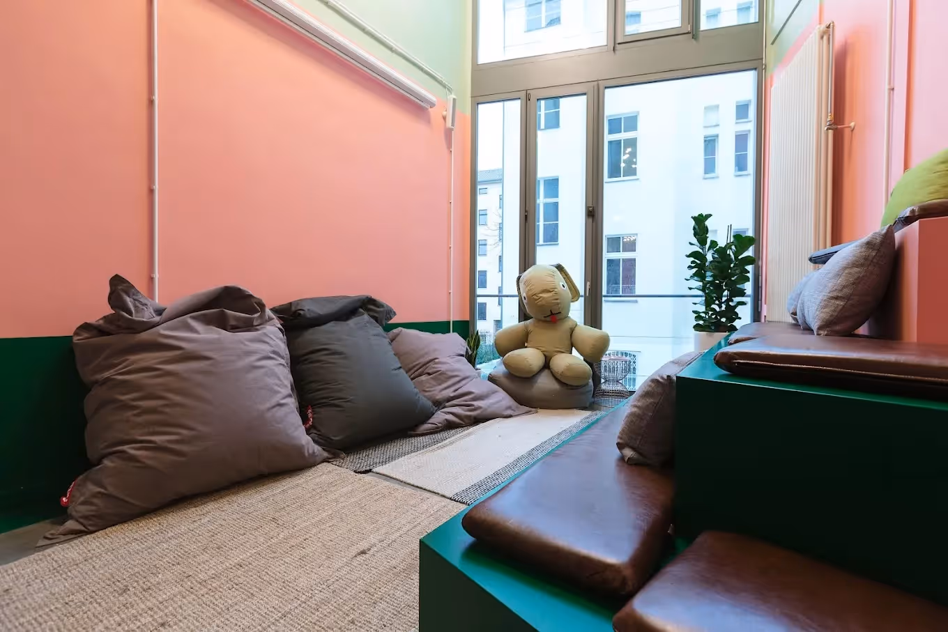Gemütlicher Raum mit Sitzsäcken, Plüschfigur, großen Fenstern, Pflanzen, bunten Wänden und Kissen auf Stufen.
