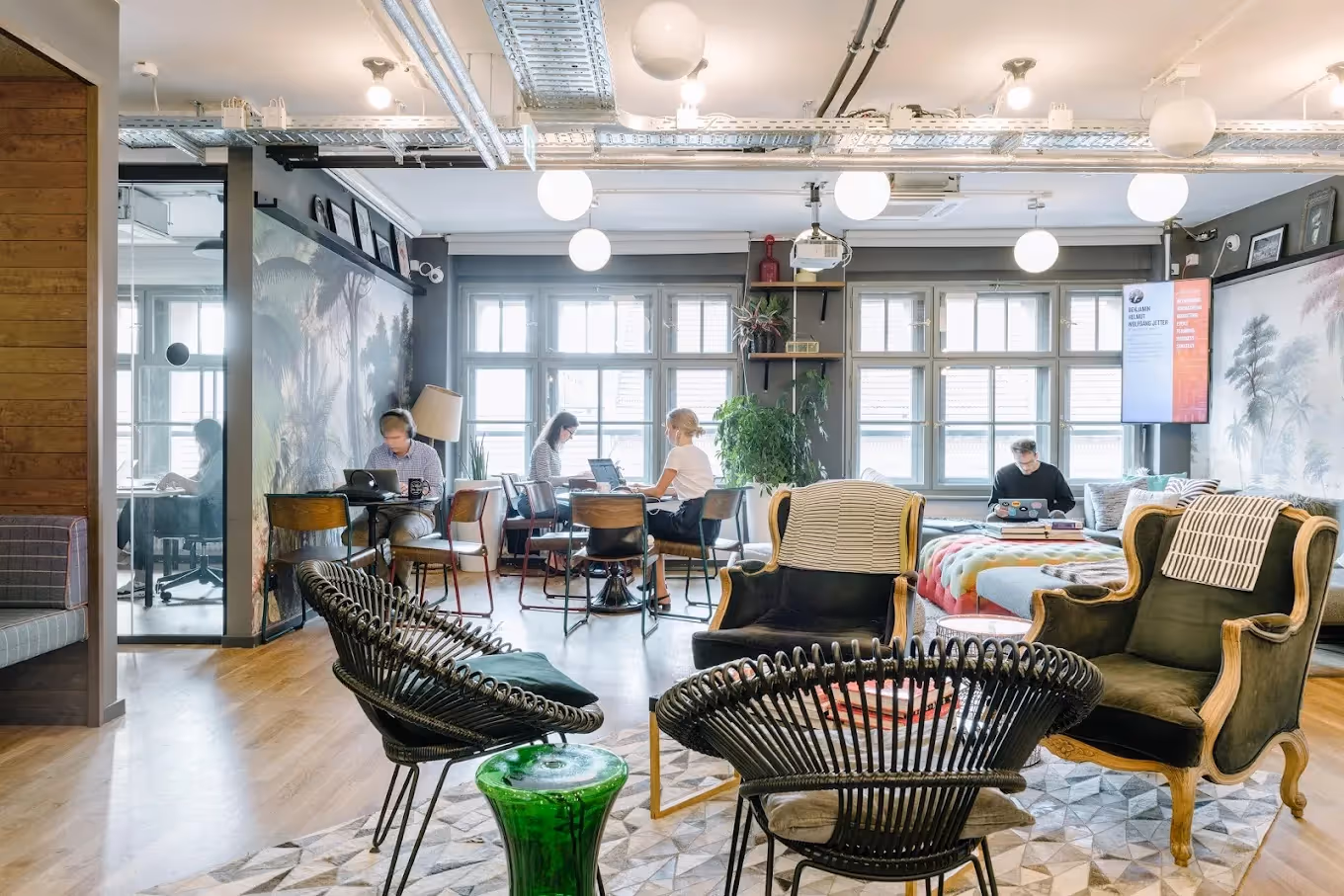 Coworking-Bereich mit Holzboden, diversen Sitzmöbeln, Pflanzen, großen Fenstern und mehreren arbeitenden Personen.