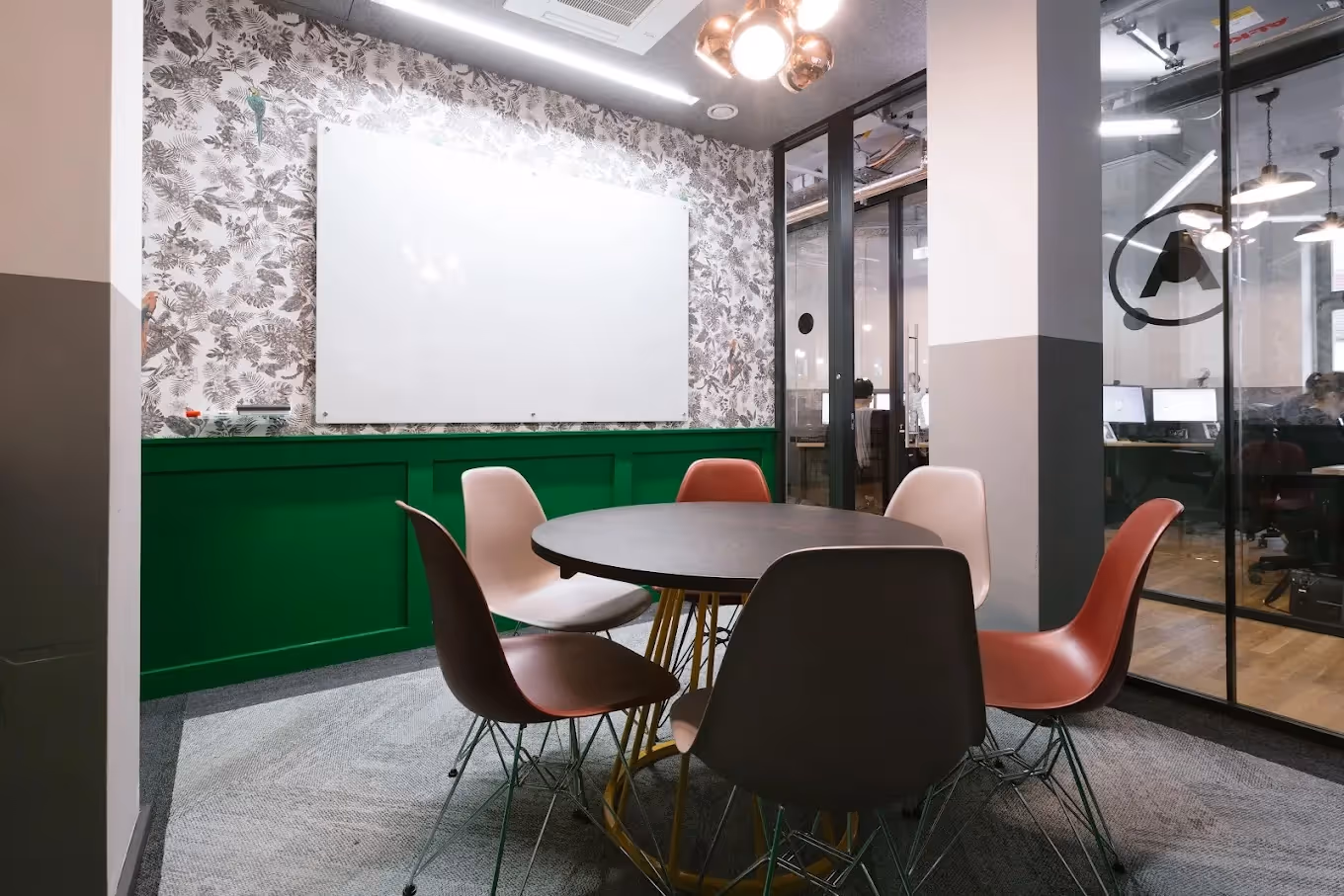 Runder Tisch mit bunten Stühlen, grüne Wandverkleidung, große Tafel, Glaswand mit Blick in Büro.