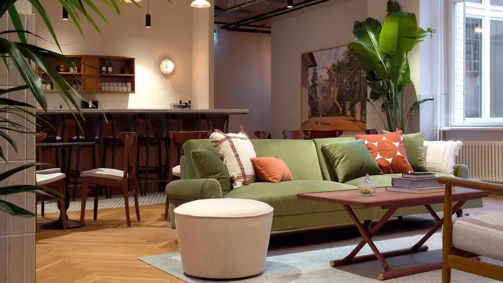 Grünes Sofa mit bunten Kissen, Holztisch, Barhocker, Pflanzen, große Fenster, hölzerner Boden, Wandbild.