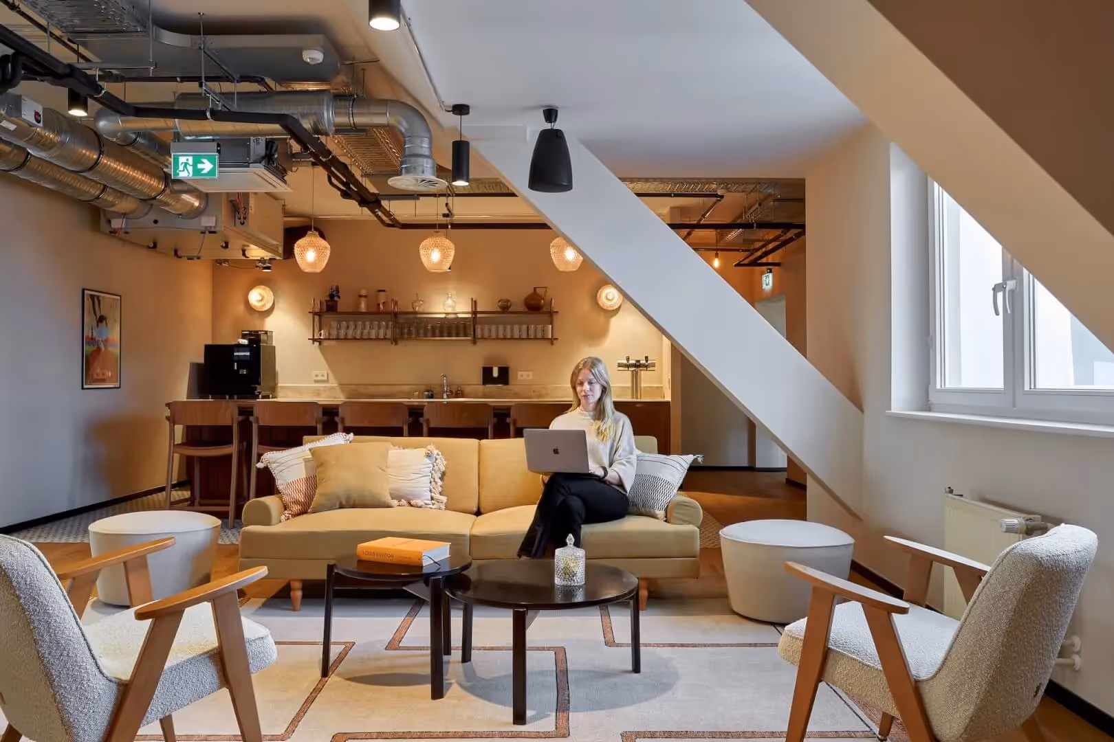Heller Raum mit Sofa, Sesseln, Couchtisch, Frau mit Laptop; Bar im Hintergrund, Hängeleuchten, große Fenster.