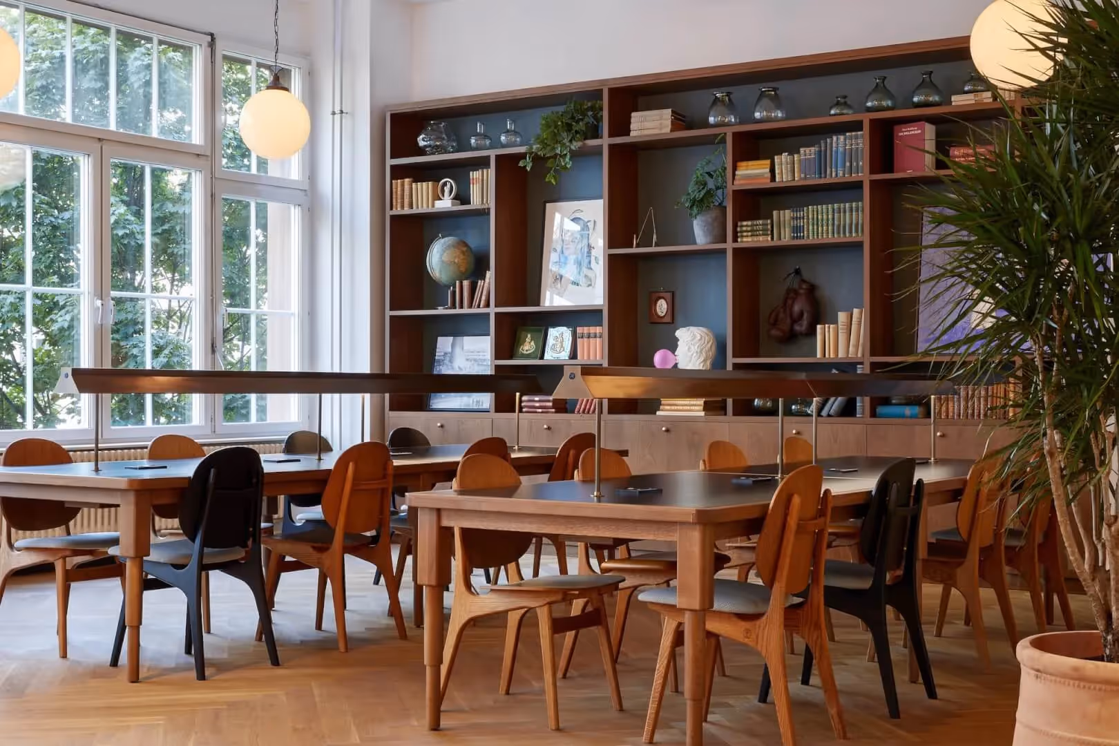 Großer Raum mit Holztischen, gemischten Stühlen, Bücherregal, Pflanzen, großen Fenstern und runden Hängelampen.