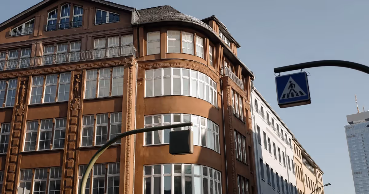 Eckgebäude mit großen Fenstern, brauner Fassade und Jugendstilelementen, Straßenschilder im Vordergrund.