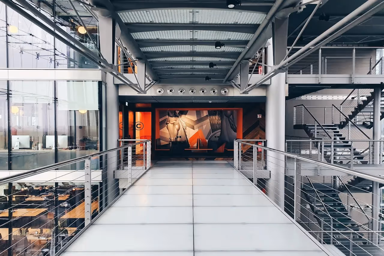 Verbindungssteg mit Glasboden, rechts Treppe, links Büroräume mit Computern, im Hintergrund bunte Wandkunst.