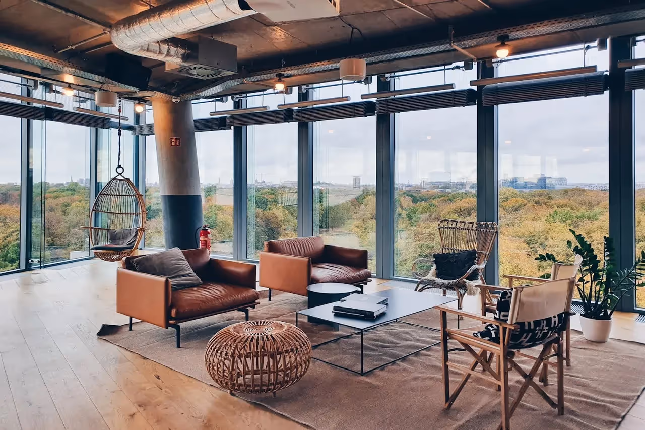 Lounge mit Ledersesseln, Hängesessel, Teppich, Glaswänden, Pflanzen, hellem Holzfußboden und Blick auf Bäume.
