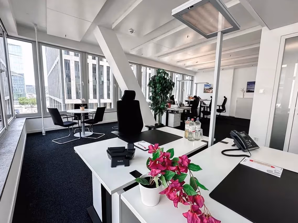 Großes Büro mit weißen Schreibtischen, schwarzen Stühlen, Pflanzen, großen Fenstern und Blick auf moderne Gebäude.