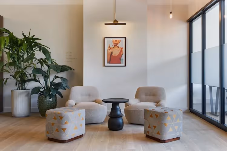 Zwei Sessel, schwarzer Tisch, zwei gemusterte Hocker, große Pflanzen, Kunstwerk an der Wand, Glaswand auf Holzfußboden.