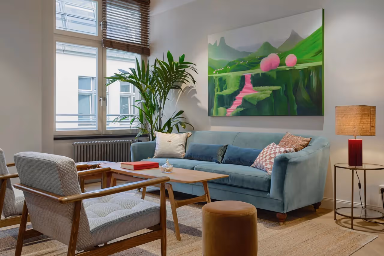 Gemütliche Sitzecke mit blauer Couch, zwei Sesseln, großem Fenster, Pflanzen, Wandbild und Tischlampe.
