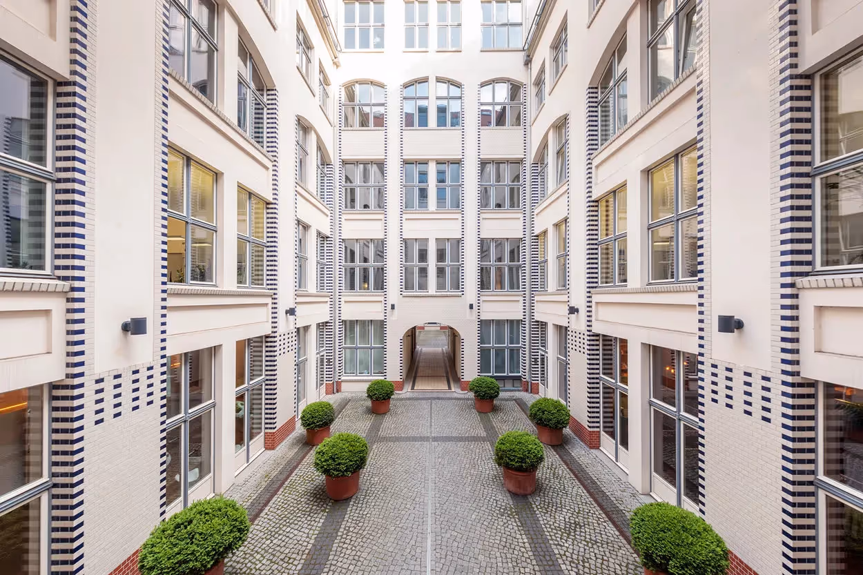 Heller Innenhof mit gepflastertem Boden, weißen Wänden, großen Fenstern und grünen Pflanzen in Töpfen.