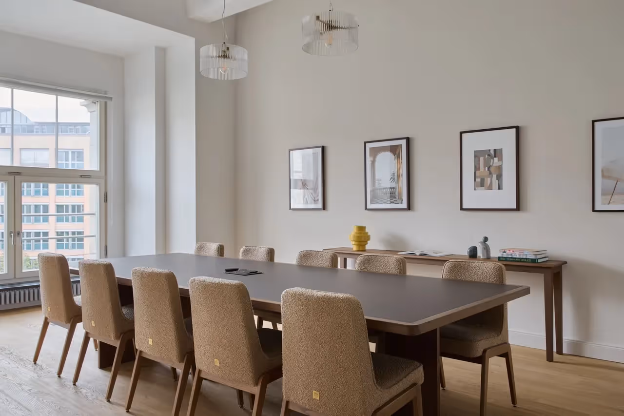 Großer Konferenztisch mit acht gepolsterten Stühlen, helle Wände mit Kunstwerken, große Fenster, Holzboden.