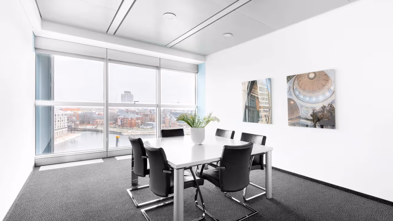 Konferenzraum mit großem Tisch, sechs schwarzen Stühlen, zwei Wandbildern und Panoramafenster mit Stadtblick.