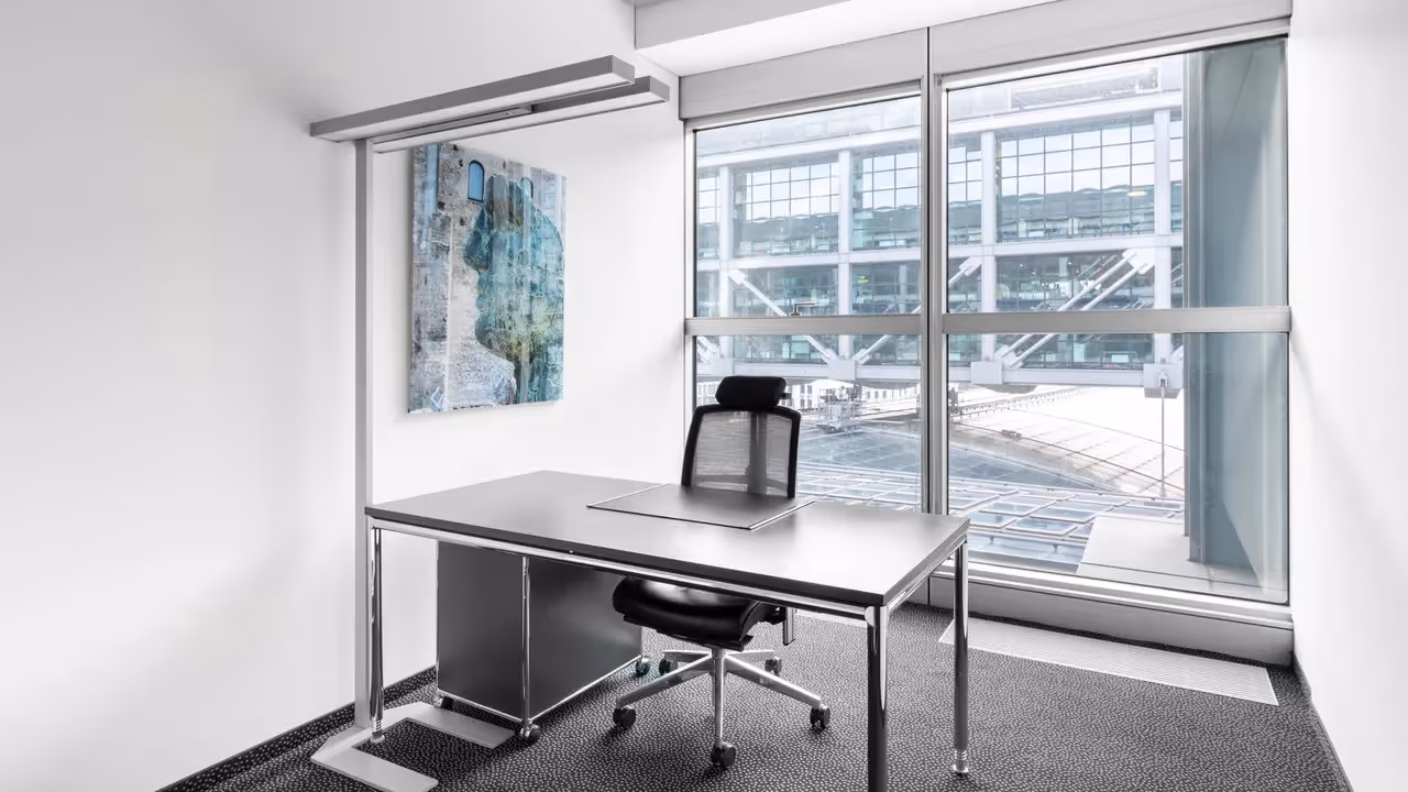 Helles Büro mit großem Fenster, modernem Schreibtisch, schwarzem Bürostuhl und Kunstwerk an der Wand.